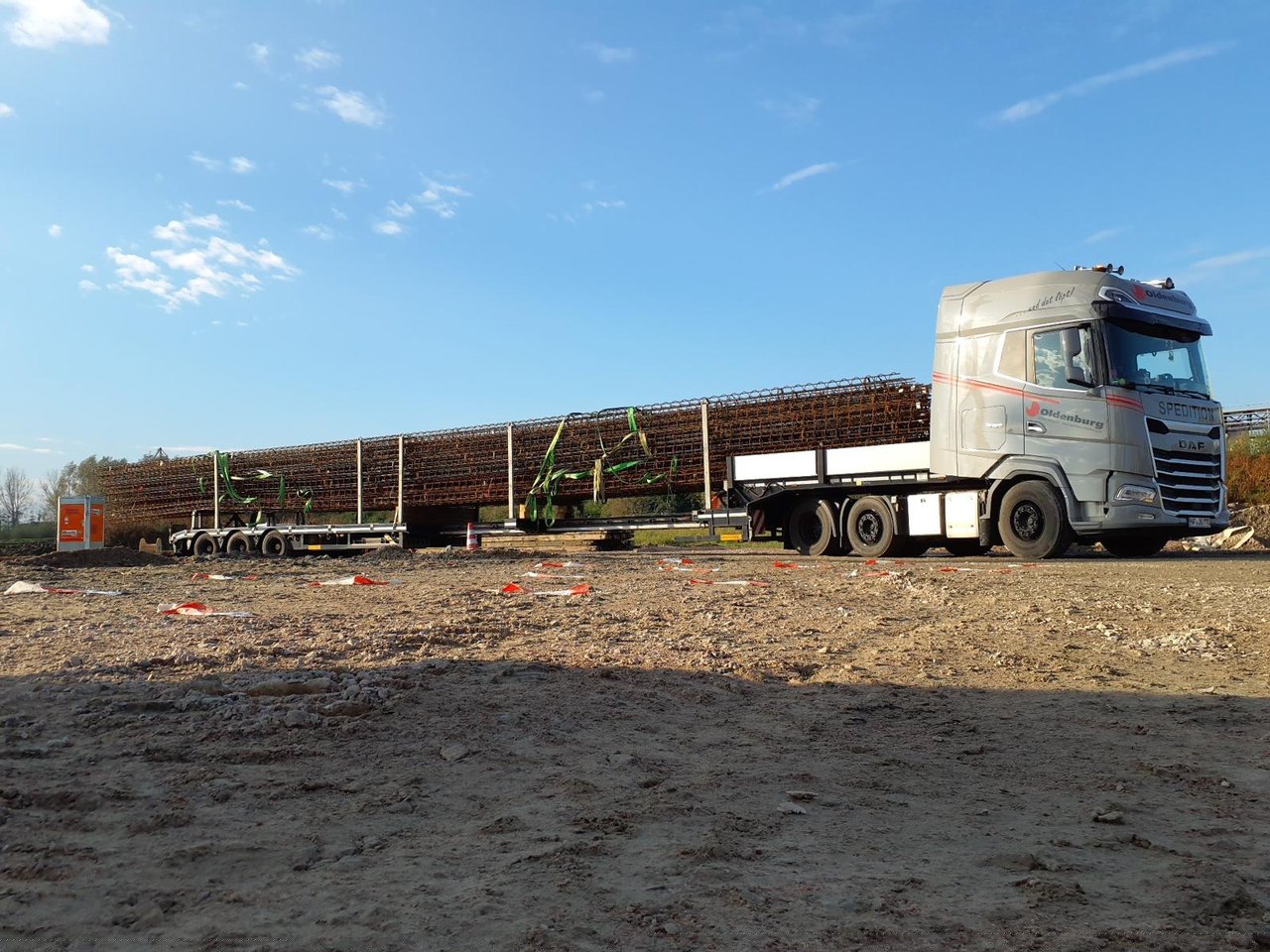 Grauer Lastwagen transportiert lange Bewehrungsstahlgitter auf einem Anhänger über eine Baustelle.